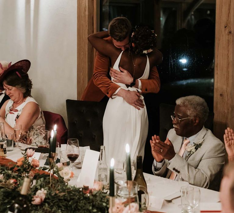 Bride in open back wedding dress with silver body chain hugging groom during wedding speeches