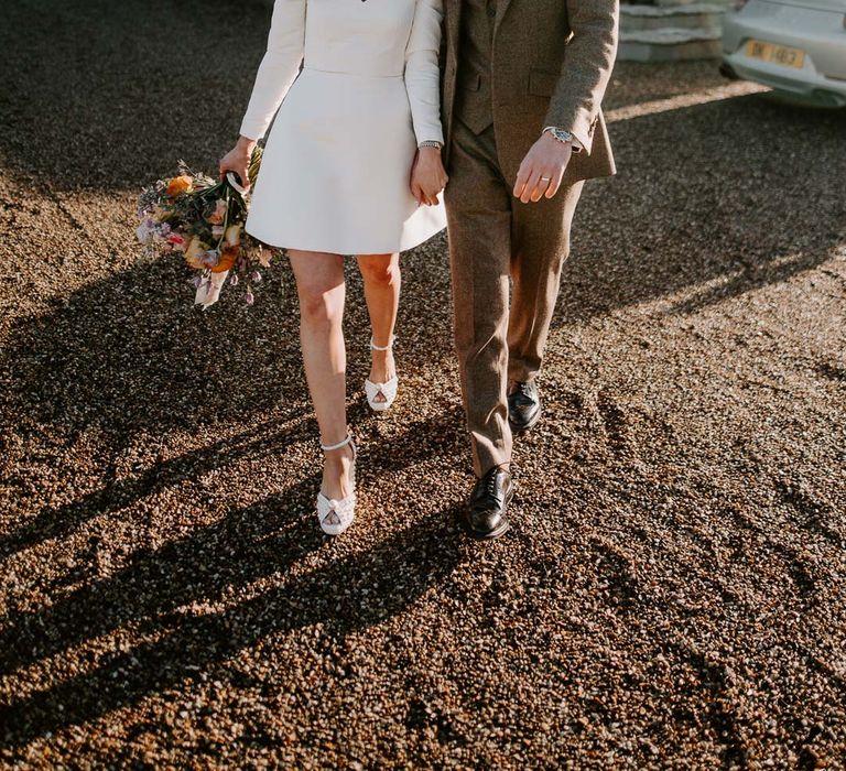 Groom in three piece chocolate brown grooms suit walking with bride in long sleeve short wedding dress, pearl bridal headband and platform pearl wedding heels at Willow Marsh Farm Loughborough 