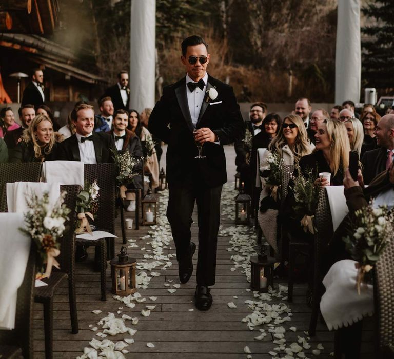 Groom in classic black tuxedo, black bowtie and white garden rose boutonniere walking down the aisle at outdoor ceremony space at Le Cabane Des Praz wedding venue with white rose petals down the aisle