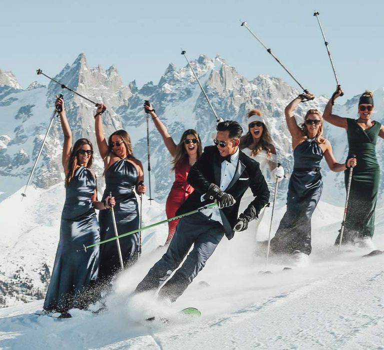 groom in classic black tuxedo, black bowtie, sunglasses and white garden rose boutonniere skiing on The French Alps with bridesmaids in satin halterneck navy bridesmaid dresses and bride in off shoulder sparkly mermaid wedding dress at French ski wedding