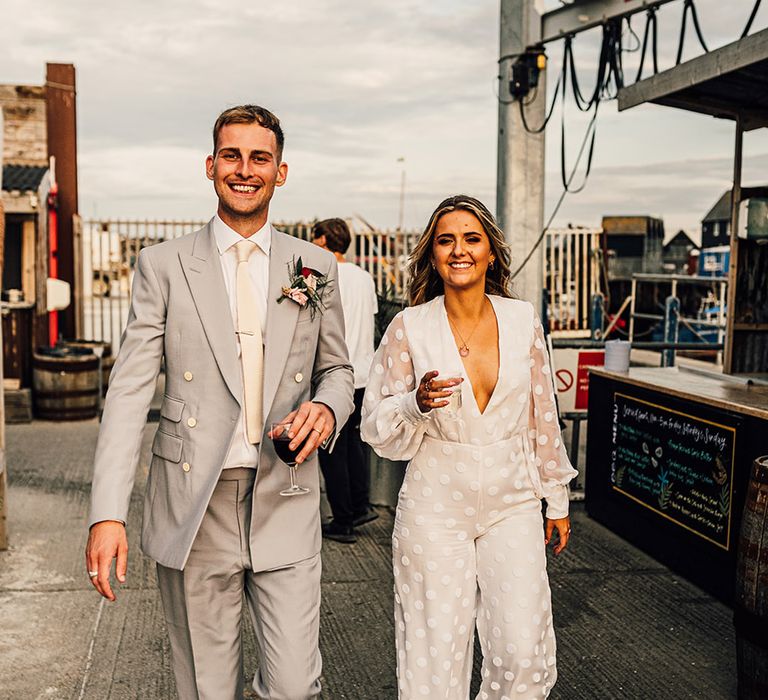 stylish bride in a wedding jumpsuit with long sleeves and groom in a light grey suit and black and white loafers at East Quay Whitstable wedding 