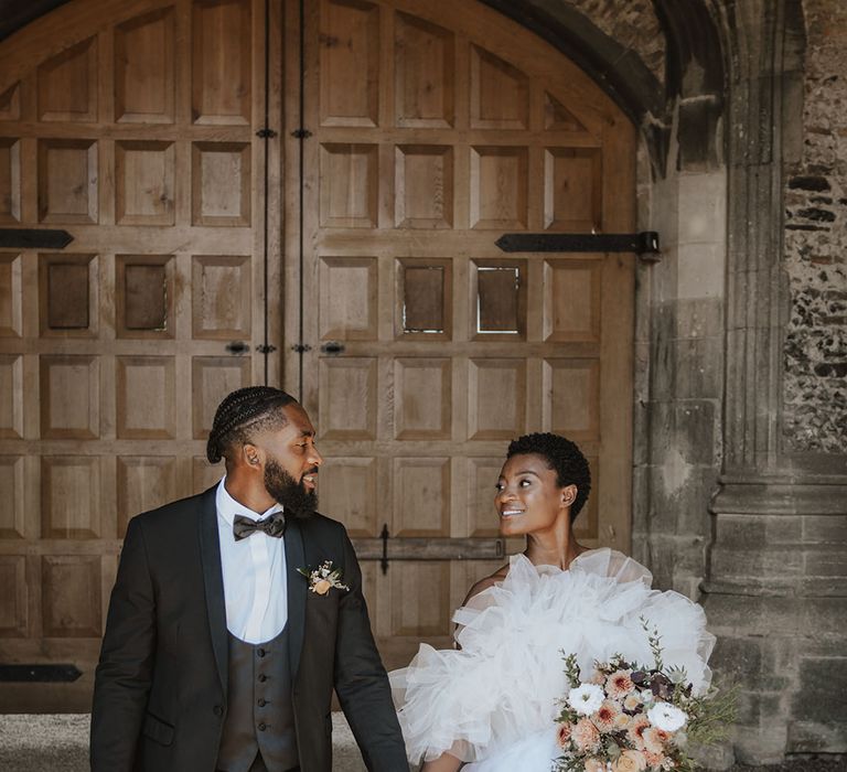 Bride walking in off the shoulder tule ruffle gown with groom in black tuxedo 