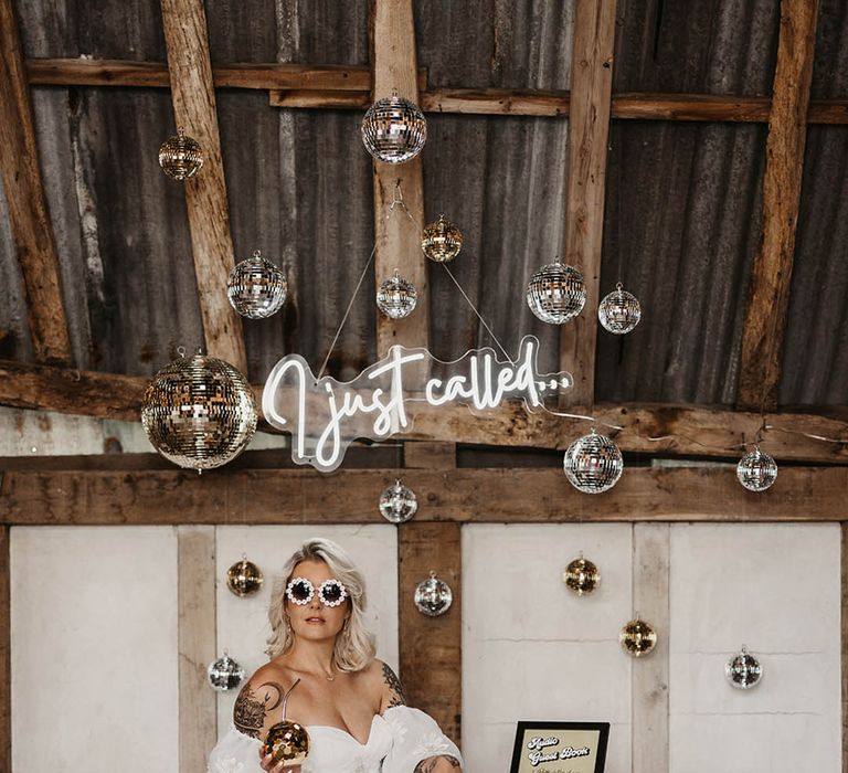 White neon wedding sign surrounded by silver and gold disco balls with bride sitting on chair wearing funky white sunglasses 