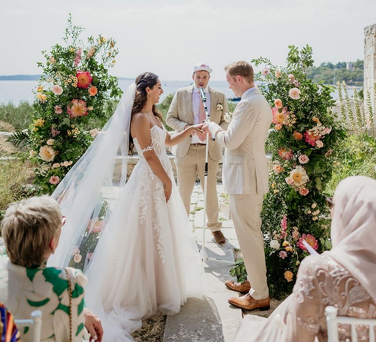 Bride in 3D lace wedding dress smiles while groom in linen suit puts on her wedding ring at altar with flower columns in Hvar, Croatia