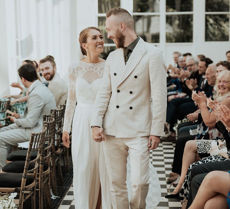 groom in a beige double breasted suit holding hands with his bride in a long sleeve wedding dress at Gunnersbury Park orangery wedding 