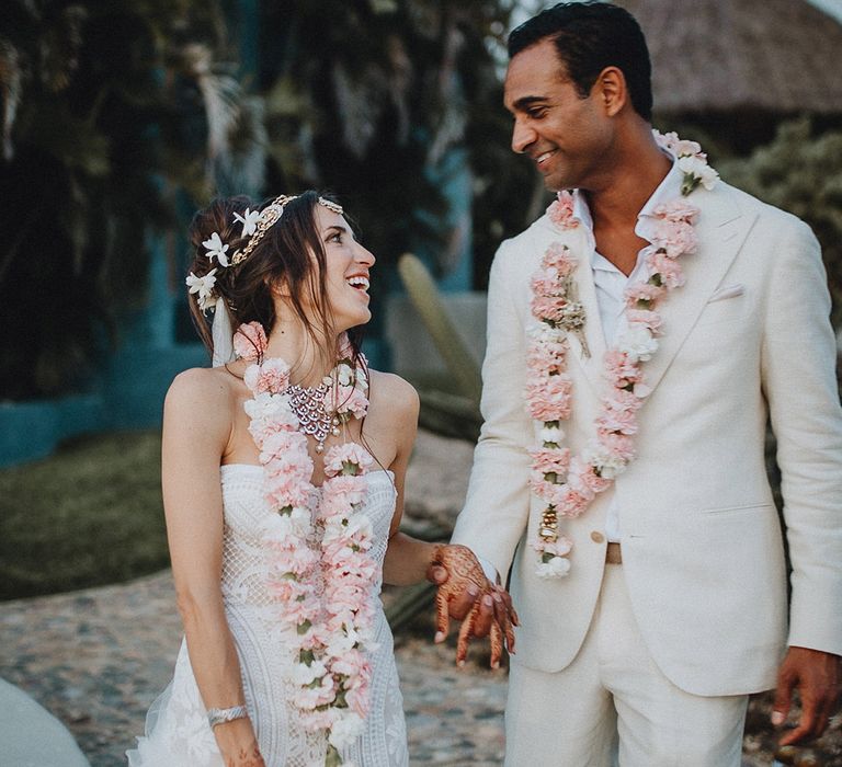 Bride and groom at boho destination wedding wearing tradtonal pink flower Varmalas. The Bride wears a lace wedding dress with flower haira ccessories and a sparkly matha patti