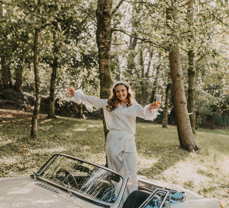 Bride in white feather wedding pyjamas standing up in the vintage cream wedding car 