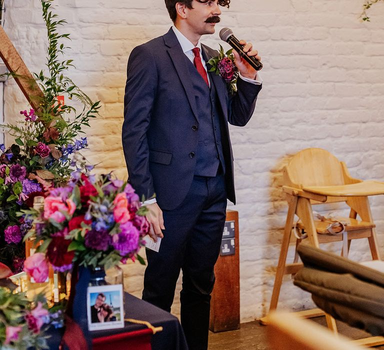 Groomsman wearing navy suit with oversized buttonhole holding microphone reading out wedding speech 