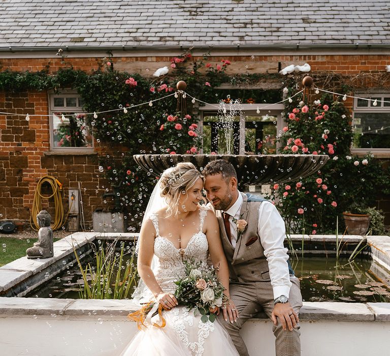 Bubbles float around the bride and groom as they sit together next to a pond 