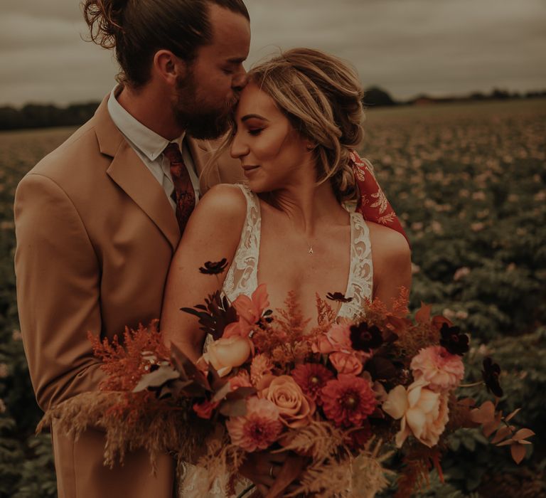 Groom in beige suit kissing his bride holding an orange flower bouquet 