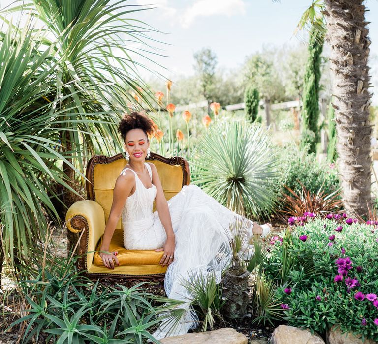 Smiling bride in Forrest by Rachel Rose Bridal dress on yellow velvet yellow armchair in botanical garden