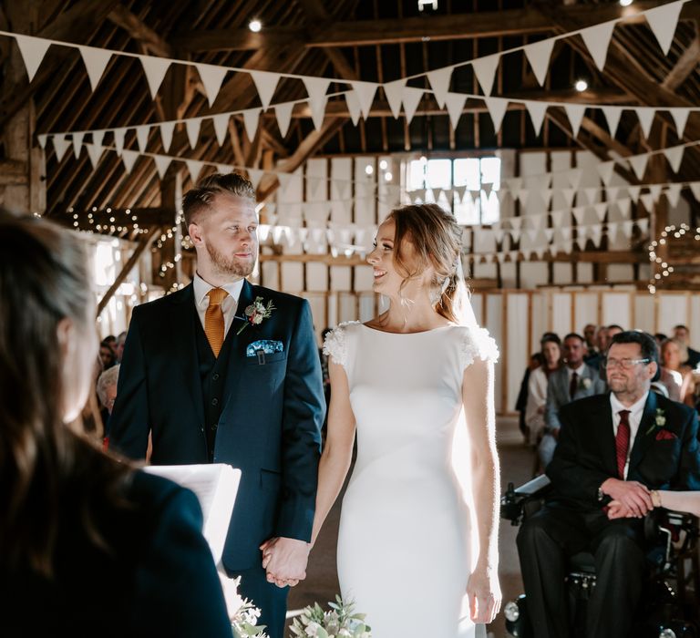 Bride in a fitted Pronovias wedding dress with applique shoulder detail standing at the altar with her groom in a navy suit and orange tie 