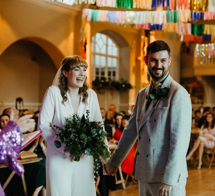 BRide in a jumpsuit holding a green foliage wedding bouquet smiling at the altar with her groom in a grey double breasted suit 