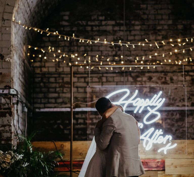 Bride and groom hugging during ceremony in front 'happily ever after' neon sign