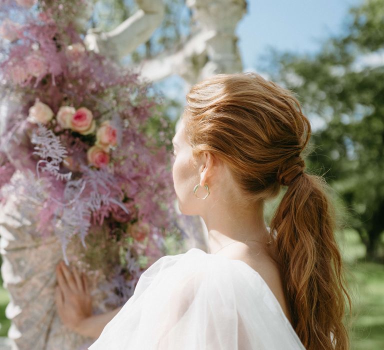 Bride wears her ginger hair in low ponytail with lilac florals in the background