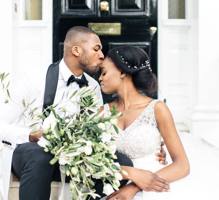 Groom kisses bride on the forehead whilst holding white floral bouquet 