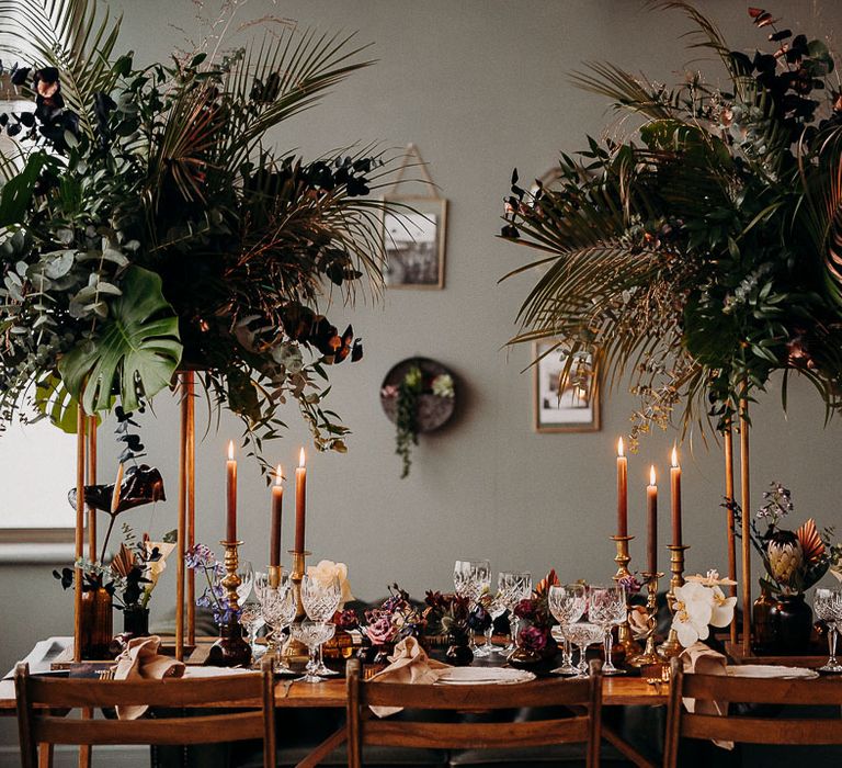 Intimate wedding table decor with high moody dark wedding flowers and foliage, candle light and opulent tableware 