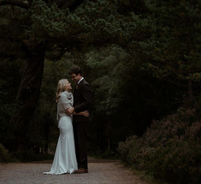 Bride wears shawl over shoulders outdoors underneath trees with groom