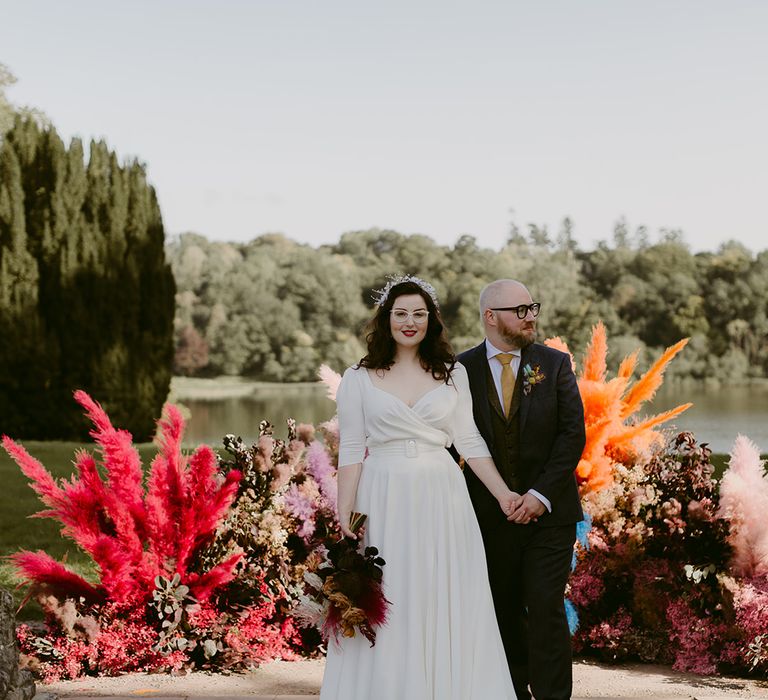 Bride in Rime Arodaky wedding dress with bridal headband holds hands with groom in navy suit and yellow tie in front of multi-coloured pampas grass floral installation by lake at Castle Leslie wedding