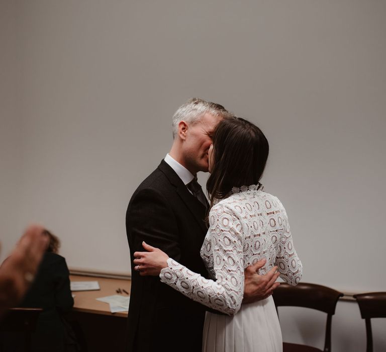 Bride in long sleeved lace top Self Portrait dress kisses groom in brown woollen blazer at Bristol Registry Office wedding