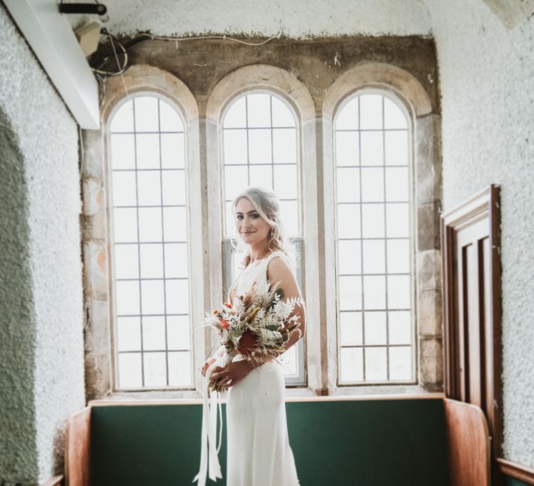 Bride stands in front of window whilst holding floral bouquet on her wedding day