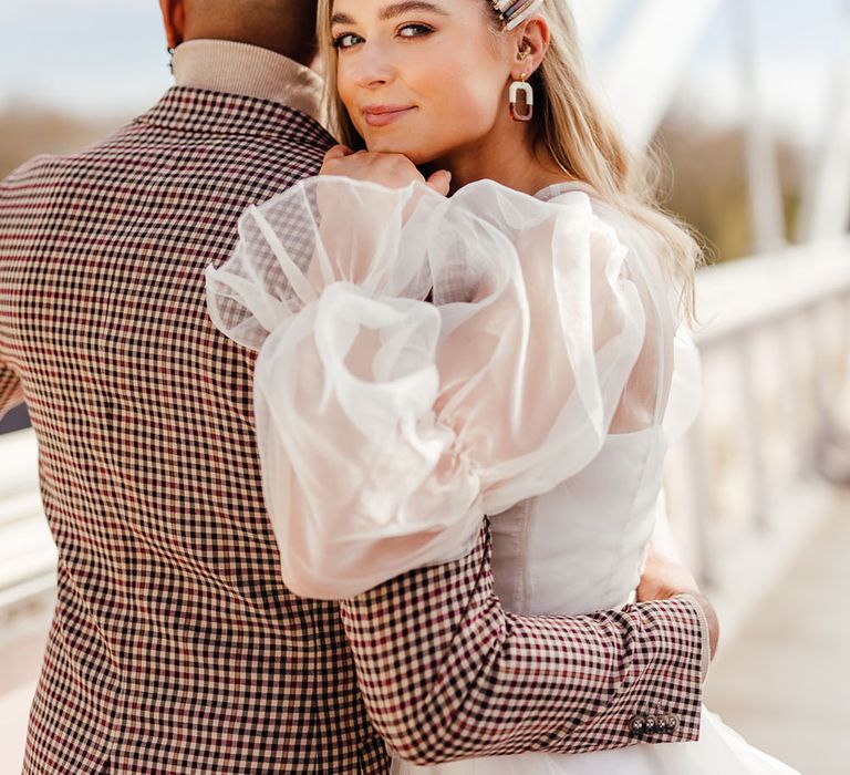 Groom in a brown check jacket with his arm around his bride in a sheer long sleeve wedding dress on Albert's Bridge in Chelsea