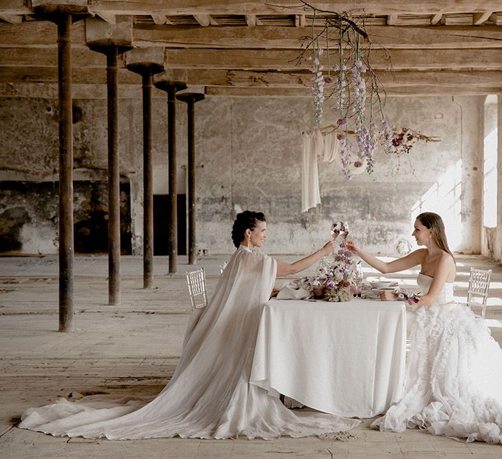 Two brides in a plain wedding cape and detachable ruffle skirt sitting at their intimate wedding reception table with purple flowers 