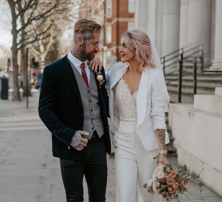 Bride in white suit with bouquet in one hand leans on the shoulder of tattooed groom in dark three piece suit