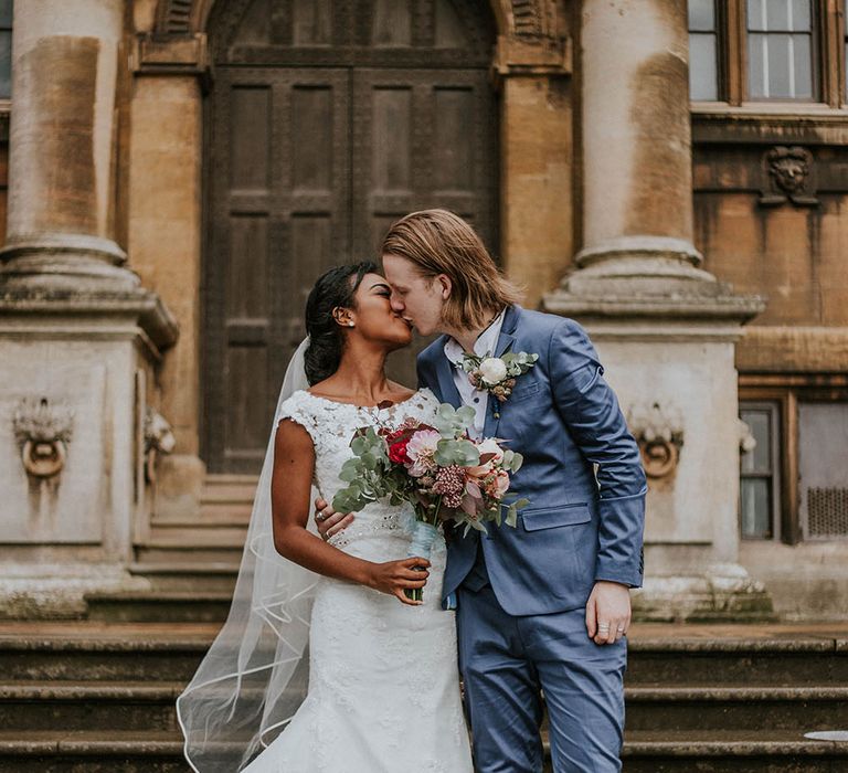 Groom in a blue suit kissing his bride in a lace wedding dress on the steps outside Heart Church in Nottingham