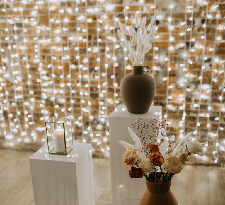 Terracotta vases with dried flowers and a rattan candle holder on pillars, in front of a wall of waterfall fairy lights