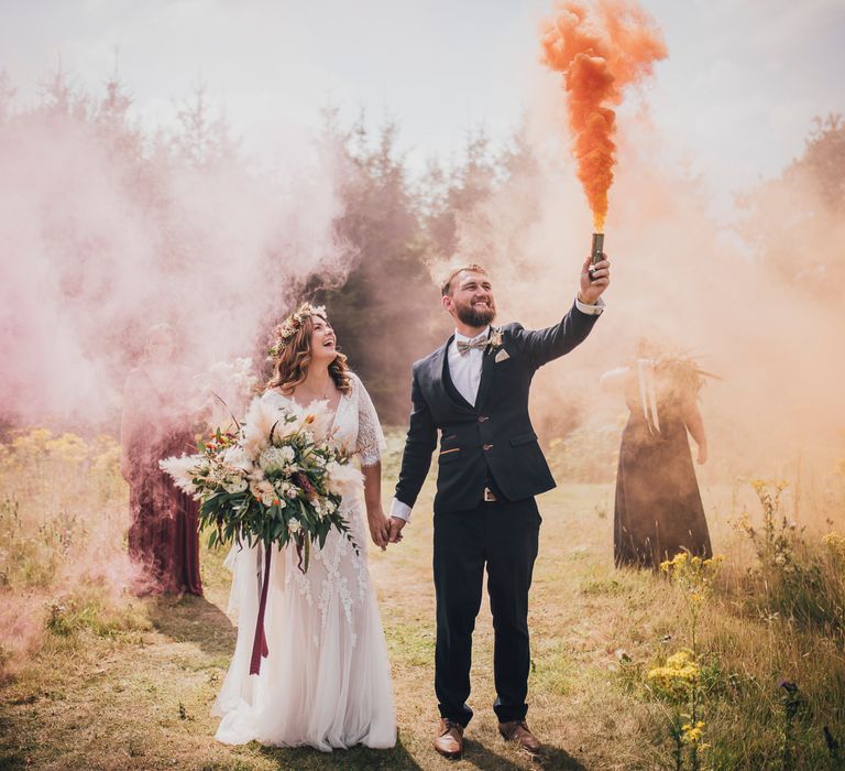 Groom holds up orange smoke bomb as he holds brides hand in field