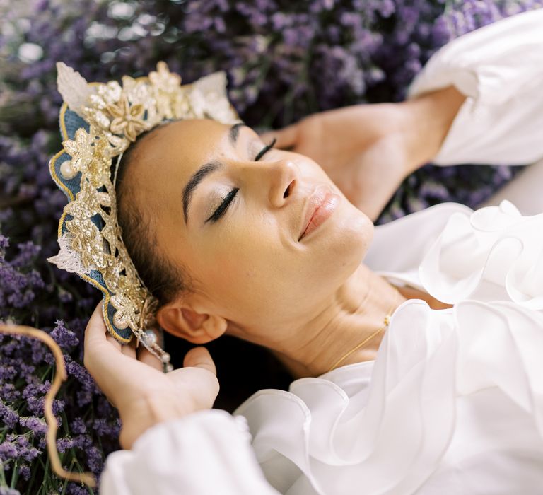 Bride lays back into lavender with eyes closed and holds golden embroidered crown