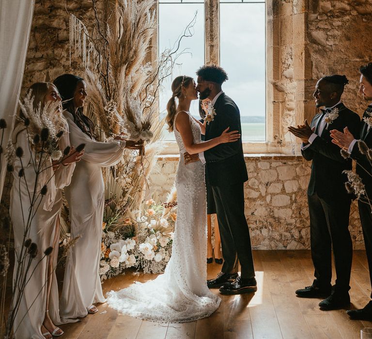 Groom in a tuxedo kissing his bride at the stone altar with window backdrop and dried and fresh flowers in neutral tones