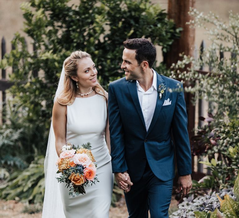 Bride & groom hold hands and walk together along the greenery as groom looks lovingly at bride