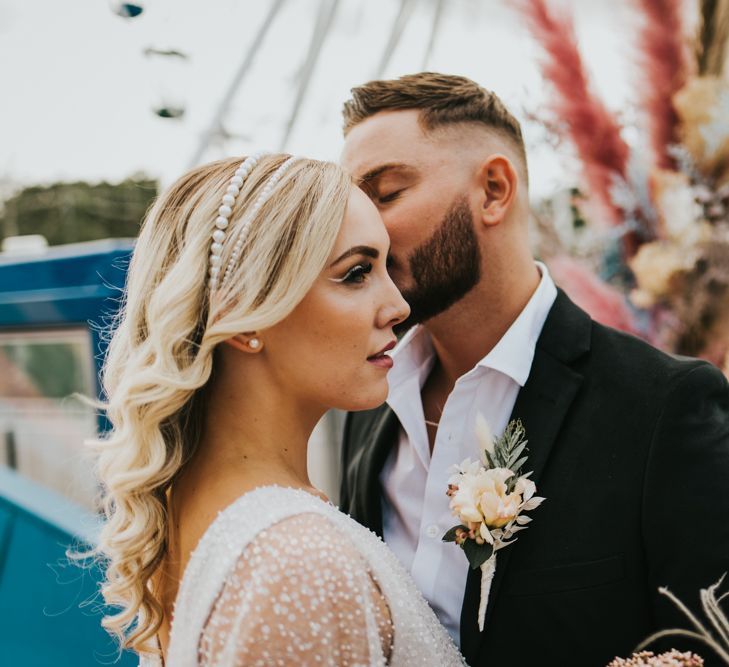 Groom in a white open collar shirt and black suit kissing his bride with long blonde wavy hair wearing a double pearl headdress