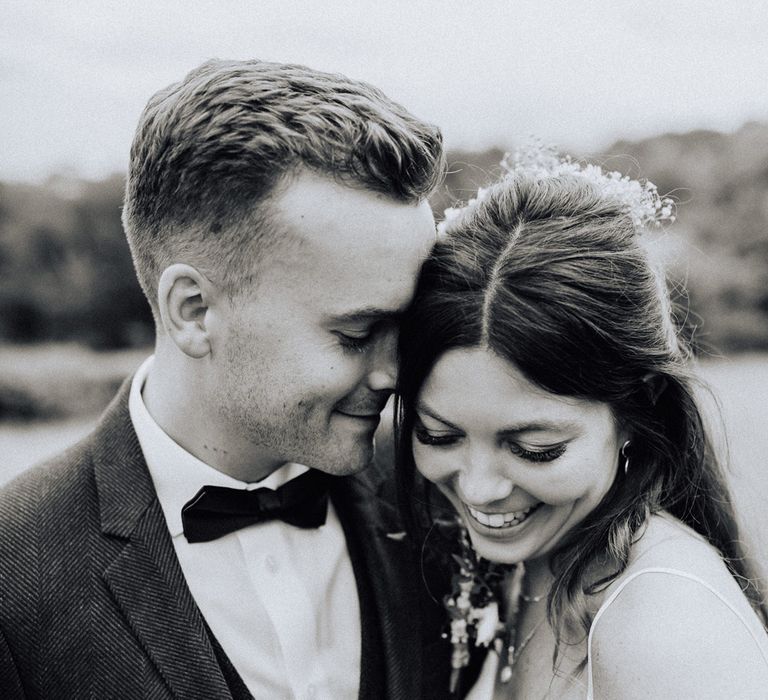 Smiling bride in white cami wedding dress and gold ribbon waistband smiles at floor whilst groom in blue suit and bow tie rests his forehead on hers at garden wedding reception