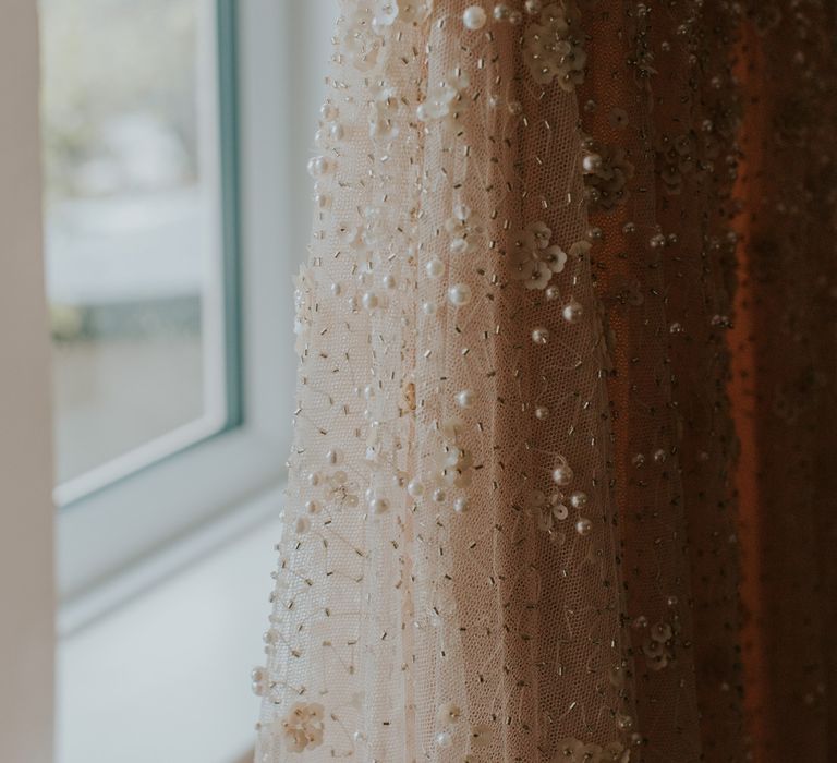 Details of pearl wedding dress with tulle and sequins hanging by window before barn wedding ceremony