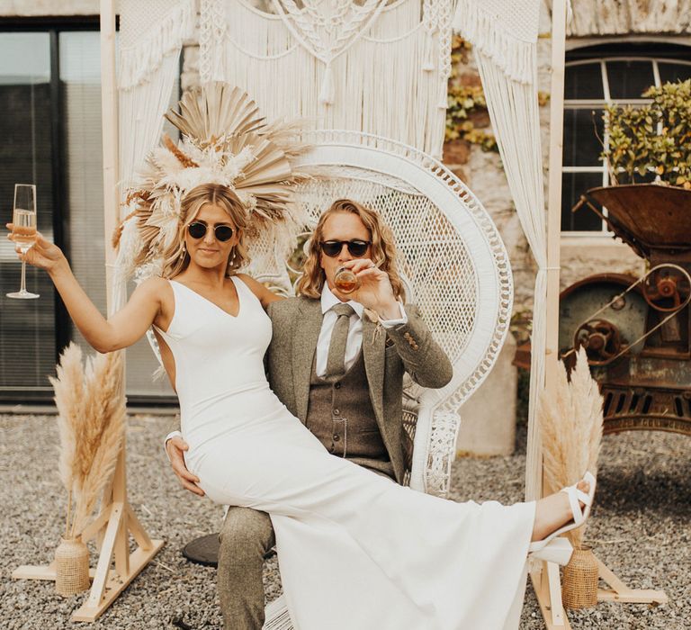 Bride in white Made With Love wedding dress and sunglasses holds up glass of sparkling wine as she sits on lap of groom in sage suit and sunglasses as he drinks beer whilst sat on white rattan throne in the courtyard of Anran Devon