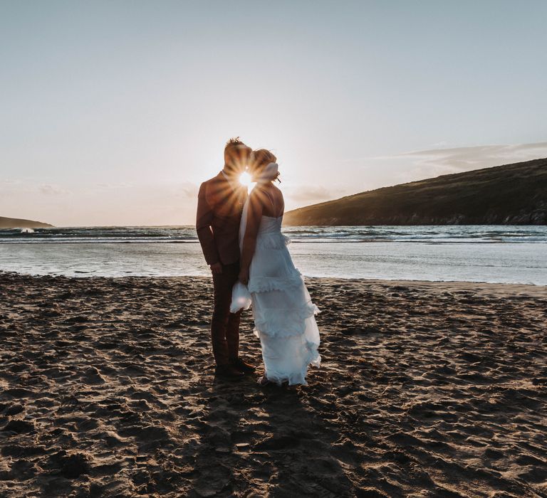 Bride & groom kiss as the sun sets behind them on the beach