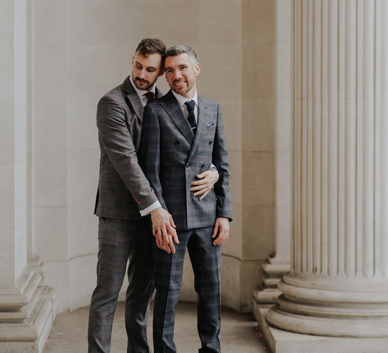 Grooms stand with one another outside Old Marylebone Town Hall in front of white pillars on their wedding day