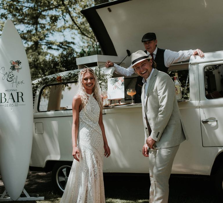 Bride in halterneck lace boho wedding dress stands with groom in linen suit and straw fedora in front of campervan cocktail bar decorated with tropical flowers for birds of paradise wedding inspiration