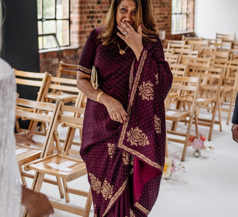 Mother of the bride in purple sari with gold detailing holds hand over mouth looking at bride in Halfpenny London wedding dress and bridal cape before wedding at Loft Studios London