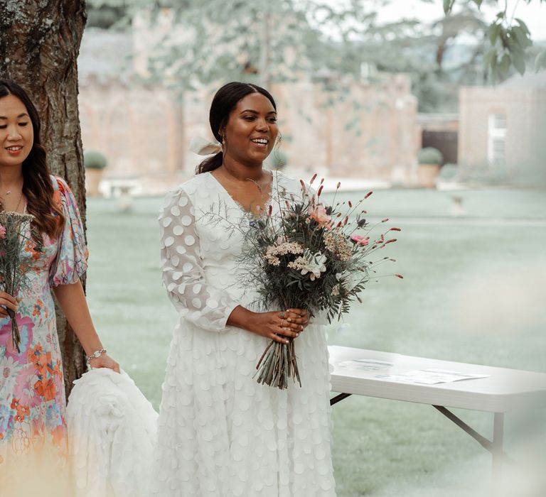 Bride in long mesh sleeved spotted Vagabond wedding dress and bridal hair bow holding mixed fresh and dried bouquet before Wasing Park wedding ceremony