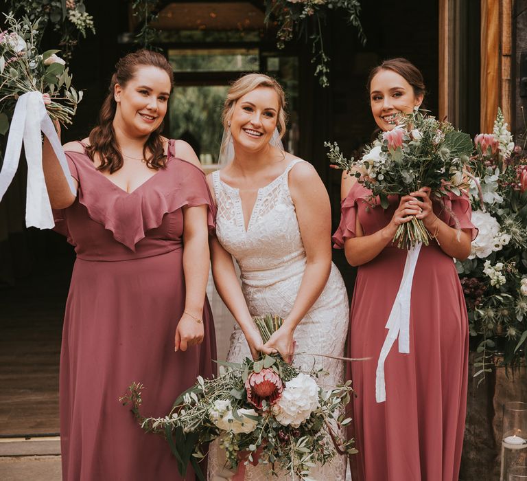 Bride in lace Justin Alexander wedding dress stands in-between two bridesmaids in dusky pink cut out shoulder bridesmaid dresses all holding pink, white and green wedding bouquets at Tythe Barn wedding with barn wedding flowers