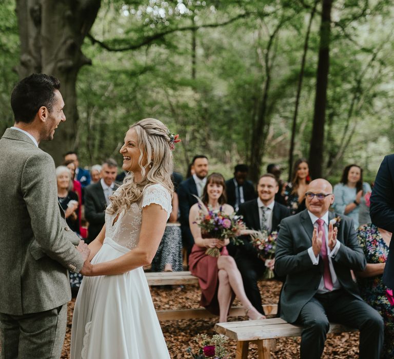 Bride in white lace cap sleeve wedding dress and flowers in her hair holds hands with groom in grey herringbone suit during woodland ceremony in Norfolk