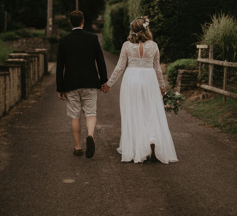 Bride and groom portrait walking down a country lane with the groom in shorts and the bride in a long sleeve wedding dress and flower crown 