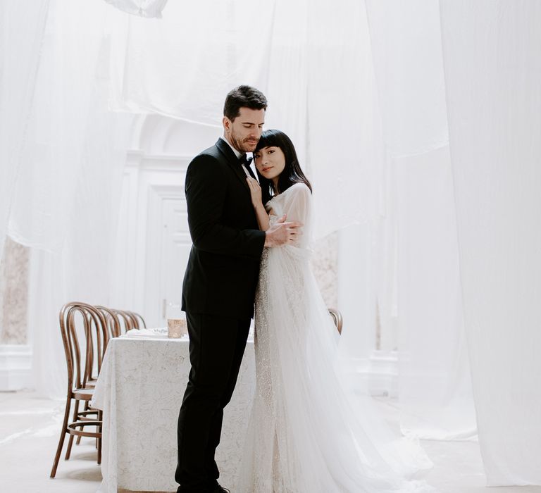 Groom in a tuxedo embracing his bride in a strapless wedding dress with embellishment under a canopy of drapes at Bylaugh Hall 