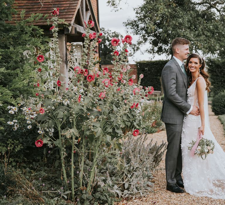 Groom in grey checked suit hugs bride in applique Berta wedding dress holding white, pink and green bridal bouquet as they stand in garden at Primrose Hill Farm for summer wedding