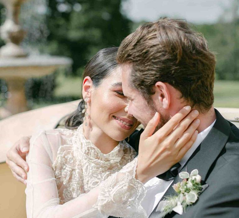 Bride in a sheer long sleeve wedding dress with high neck design and lace cuffs embracing her grooms face in the back of a vintage wedding car 