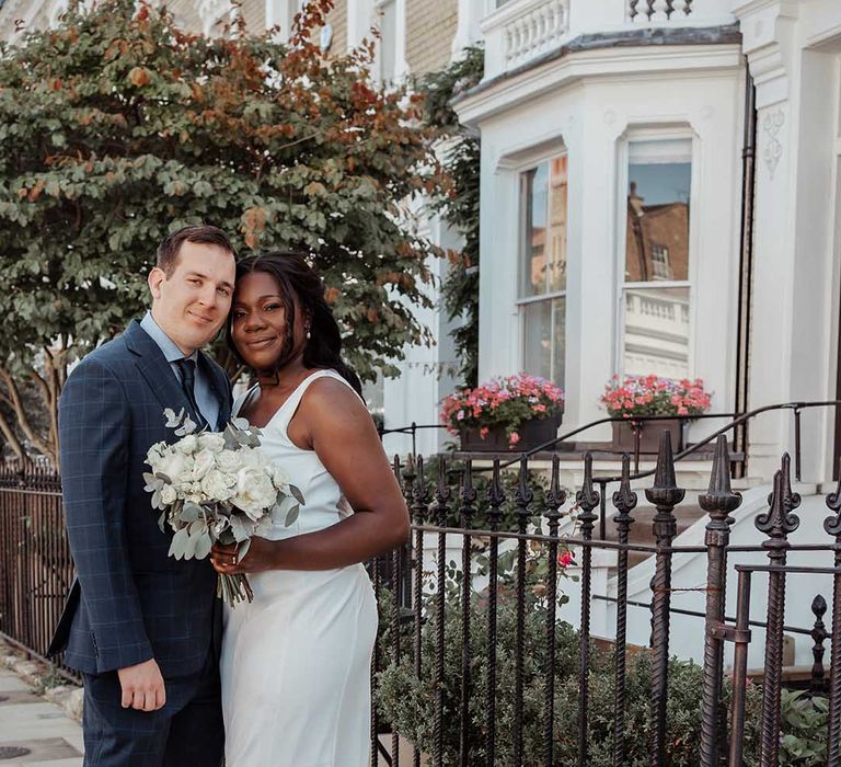 Bride & groom stand outside in Chelsea on their wedding day as bride holds classic white floral bouquet